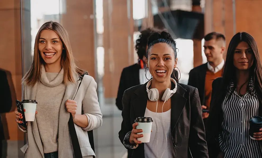 Personer som går tillsammans i en modern inomhusmiljö och håller med kaffemuggar, med kontors- eller företagsmiljö i bakgrunden.