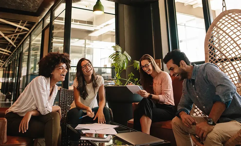 Personer i ett mötesutrymme med glasväggar, sitter i soffor och stolar och går igenom dokument i en informell arbetsmiljö.