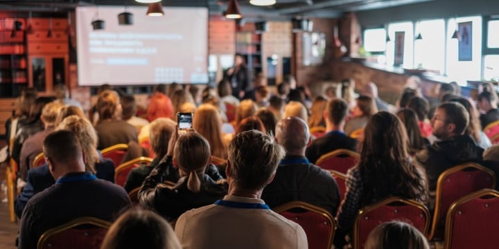 Publik sitter i en konferenslokal och lyssnar på en presentation på scen.