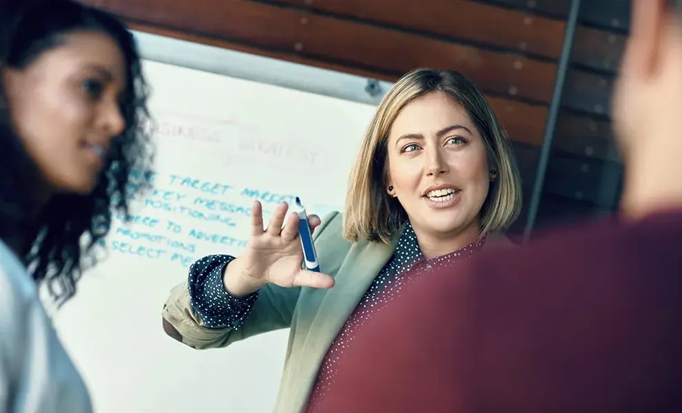 En person håller en presentation framför en grupp och gestikulerar med en penna vid en whiteboard med anteckningar i bakgrunden.
